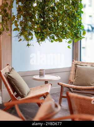 Gemütliche Café-Ecke mit Rattanstuhl, kleinem runden Tisch mit Kaffee, Gebäck und einer Vase mit getrockneten Blumen, umgeben von üppigen hängenden Pflanzen Stockfoto