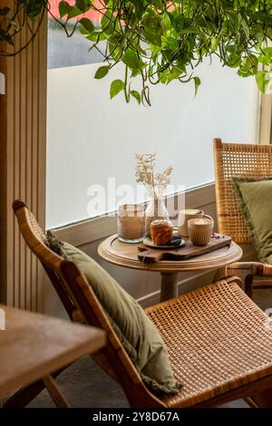 Gemütliche Café-Ecke mit Rattanstuhl, kleinem runden Tisch mit Kaffee, Gebäck und einer Vase mit getrockneten Blumen, umgeben von üppigen hängenden Pflanzen Stockfoto