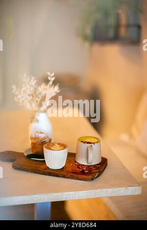 Gemütlicher Café-Tisch mit zwei Kaffeetassen aus Keramik, einem Gebäck, einer weißen Vase mit getrockneten Blumen und einem Magazin, das in warmem natürlichem Licht getaucht ist. Stockfoto