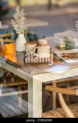 Gemütlicher Café-Tisch mit zwei Kaffeetassen aus Keramik, einem Gebäck, einer weißen Vase mit getrockneten Blumen und einem Magazin, das in warmem natürlichem Licht getaucht ist. Stockfoto