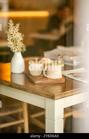 Gemütlicher Café-Tisch mit zwei Kaffeetassen aus Keramik, einem Gebäck, einer weißen Vase mit getrockneten Blumen und einem Magazin, das in warmem natürlichem Licht getaucht ist. Stockfoto