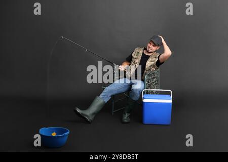 Fischer mit Rute und Kühlbox Angeln von Schüssel auf Stuhl vor schwarzem Hintergrund Stockfoto