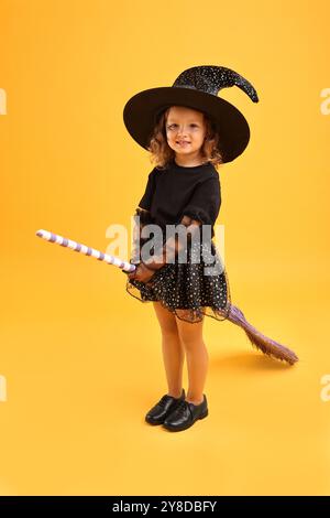 Süßes Mädchen mit Besen, verkleidet wie Hexe auf gelbem Hintergrund. Halloween-Feier Stockfoto