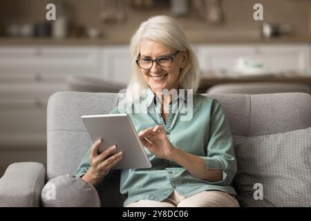 Lächelnde ältere Frau, die auf der Couch sitzt und Tablette benutzt Stockfoto