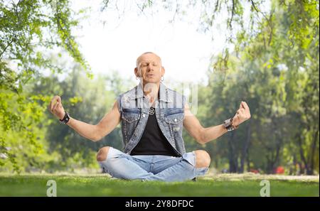 Glatzkopf in Jeans sitzt mit überkreuzten Beinen und übt Meditation in einem Park Stockfoto