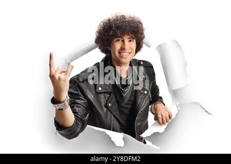 Junger Mann in einer Lederjacke, der durch ein Papierloch schaut und ein Rock-and-Roll-Schild auf weißem Hintergrund mit Gesten zeigt Stockfoto