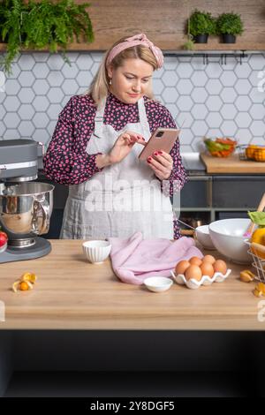 In einer gemütlichen Heimküche prüft der Bäcker das Rezept auf dem Smartphone, während er von Zutaten und Geräten im Küchenkonzept umgeben ist Stockfoto