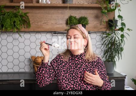 In einer gemütlichen Küche, fröhliche Frau, die Joghurt genießt, eine Blume und ein Stirnband trägt, umgeben von Zimmerpflanzen, Glücks- und Genusskonzept Stockfoto