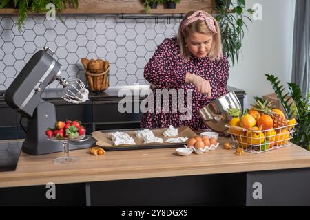 In der Küche peitscht eine Frau Meringue mit einem elektrischen Mixer, mit frischem Obst und Zutaten auf der Theke, Hausbackkonzept Stockfoto