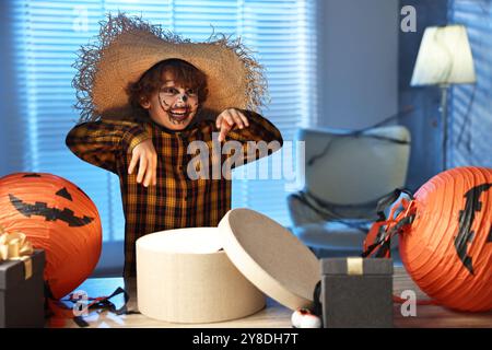 Süßer Junge, der wie Vogelscheuche gekleidet ist, mit festlichem Dekor und Geschenkboxen drinnen in der Nacht. Halloween-Feier Stockfoto