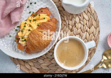 Ein Teller mit einem Croissant-Sandwich und einer Seite Zwiebeln. Der Teller wird mit einer Tasse Kaffee auf einen Tisch gestellt. Die Szene ist lässig und einladend Stockfoto