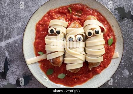 Drei Hot Dogs mit gezeichneten Augen sitzen auf einem Teller Spaghetti mit roter Sauce. Der Teller ist mit einem gruseligen Halloween-Thema dekoriert, Addin Stockfoto