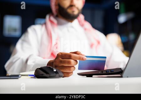 Selektiver Fokus auf einen Mann aus dem Nahen Osten, der eine Debitkarte für nahtloses Online-Shopping ergreift. Nahaufnahme eines arabischen Mannes in traditioneller Kleidung, der mit seinem Computer bezahlt. Stockfoto