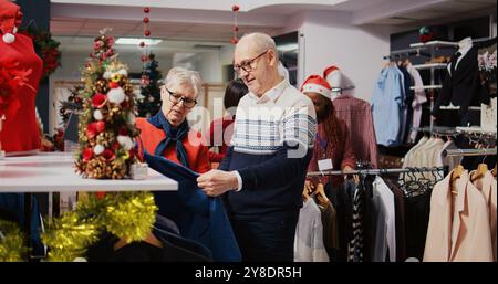 Ältere Ehemann und Frau stöbern während der Weihnachtseinkäufe durch die Kleiderständer im Weihnachtsgeschäft. Ältere Kunden, die formelle Kleidung kaufen möchten und sich an Werbeaktionen für Feiertage freuen Stockfoto