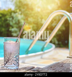 Ein Cocktailglas auf einer Holzterrasse am türkisfarbenen Pool mit Edelstahlleiter und mediterranen Bodenfliesen in der Sommersonne Stockfoto