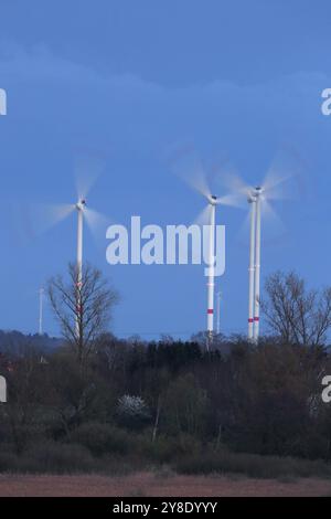 Leuchtende Windräder rotieren in einem von Bäumen umgebenen Bereich im Abendlicht, Windräder im Abendlicht, lange Exposition, Naturpark flus Stockfoto