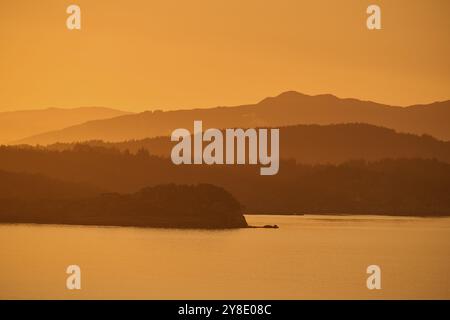 Ein sanfter oranger Sonnenaufgang hinter den dunklen Silhouetten der Berge und eines ruhigen Meeres, Herbst, Bergen, Nordsee, Norwegen, Europa Stockfoto