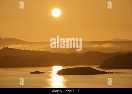 Ein sanfter oranger Sonnenaufgang hinter den dunklen Silhouetten der Berge und ein ruhiges Meer, Sonnenstrahlen spiegeln sich in der Nordsee, im Herbst, Bergen, Nordsee, Norwegen Stockfoto