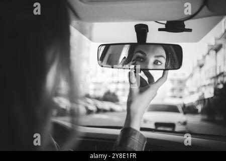 Junge Frau im Rückspiegel auf der Suche und im Auto machen. Moderne geschäftigen Leben. schöne Mädchen sitzen im Fahrersitz des Autos tun, Make-up, Appl Stockfoto