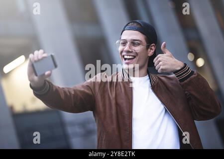 Selfie-Manie der junge Mann macht Selfie mit der Kamera. Er trägt lässige, trendige Kleidung und einen Schnappverschluss Stockfoto