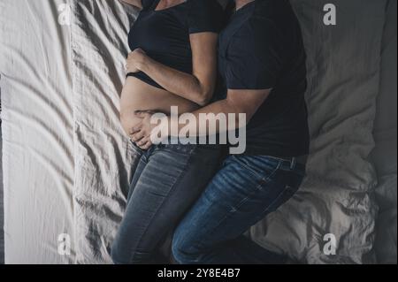 Ein Paar liegt im Bett, der Mann umschließt die schwangere Frau und legt seine Hand auf den Bauch des Babys Stockfoto