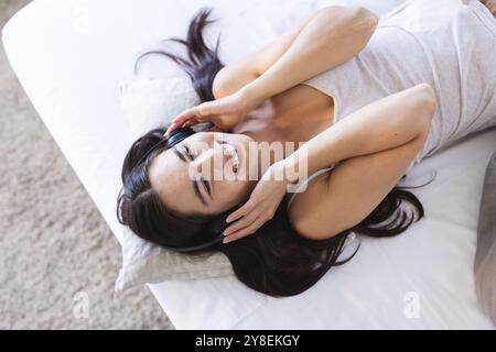 Hochwinkelansicht einer lächelnden jungen Frau aus kaukasien, die Musik über Kopfhörer hört, während sie auf dem Bett liegt Stockfoto