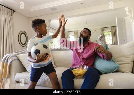 Glücklicher afroamerikanischer Vater und Sohn, der auf dem Sofa sitzt, Fußballspiele ansieht und Chips isst Stockfoto