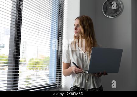 Vorderansicht Nahaufnahme eines jungen kaukasischen stehende Frau mit einem Laptop mit den Kopf durch ein Fenster mit Jalousien drehte sich in Stockfoto