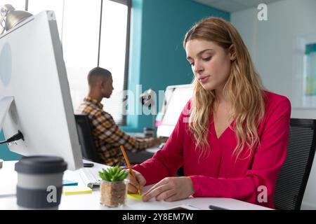 Vorderansicht Nahaufnahme eines jungen kaukasischen Frau sitzt am Schreibtisch schreiben eine Notiz, mit ihren männlichen Kollegen sitzen und an einem Computer in der Rückseite arbeiten Stockfoto