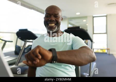 afroamerikaner Senior-Mann, der eine Smartwatch benutzt, während er im Fitnessstudio auf dem Heimtrainer sitzt Stockfoto