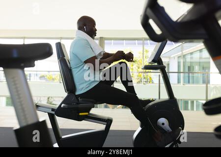 afroamerikanischer Senior-Mann, der eine Smartwatch benutzt, während er im Fitnessstudio auf dem Heimtrainer trainiert Stockfoto