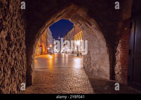 Blick durch das Florianska-Tor in Krakau, Polen, zeigt die kopfsteingepflasterte Straße, die nachts in Richtung Marienkirche führt. Das historische Tor und die Straße sind mit Straßenlaternen beleuchtet. Stockfoto