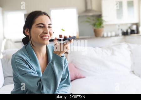 Lächelnde weiße junge Frau, die über Lautsprecher über das Smartphone spricht, während sie zu Hause auf dem Sofa sitzt Stockfoto