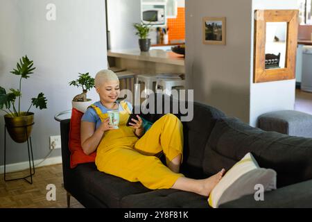 Glückliche modische unveränderte birassische Frau, die sich zu Hause auf der Couch mit Kaffee entspannt, mit Smartphone Stockfoto