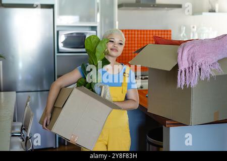 Glückliche, modische, unveränderte Frau, die Pflanzen in Kisten trägt, Besitztümer verpackt, um das Haus umzuziehen Stockfoto