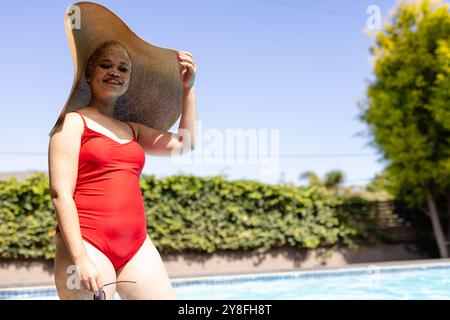 Glückliche Frau in Badeanzug und Sonnenhut am Pool, lächelnd im Sonnenlicht, Kopierraum Stockfoto