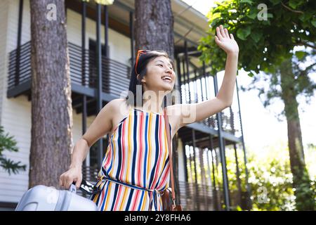 Glückliche asiatische Frau in farbenfrohem Kleid mit der Hand winkenden Koffern im Garten zu Hause Stockfoto