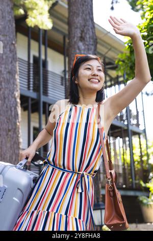 Glückliche asiatische Frau in farbenfrohem Kleid mit der Hand winkenden Koffern im Garten zu Hause Stockfoto