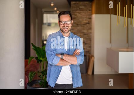 Porträt eines erfolgreichen jungen Geschäftsmannes mit überkreuzten Armen und Brillen im modernen Büro Stockfoto