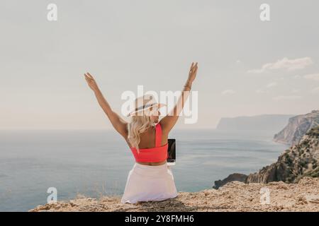 Eine Frau sitzt auf einem Hügel mit Blick auf das Meer mit einem Laptop im Schoß. Sie trägt ein rosafarbenes Oberteil und einen Strohhut. Konzept der Entspannung und Stockfoto