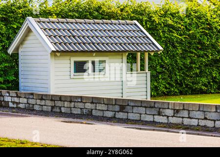 Kleiner weißer Schuppen mit gekacheltem Dach neben Betonmauer in Wohngebiet, umgeben von grüner Hecke. Schweden. Stockfoto