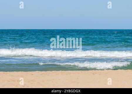 Dieses Bild zeigt eine ruhige Strandszene mit klarem blauem Himmel, einem ruhigen Ozean mit sanften Wellen, die auf das Sandufer brechen. T Stockfoto