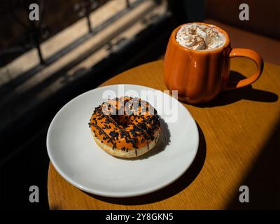 Herbst Kürbis Gewürz Latte in einem orangen Kürbisbecher mit Schlagsahne und Zimt, orange glasierter Donut Stockfoto