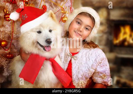 Porträt eines kleinen Mädchens, das in einer Umarmung mit dem Samoiden-Hund im Hintergrund des Weihnachtsbaums sitzt Stockfoto