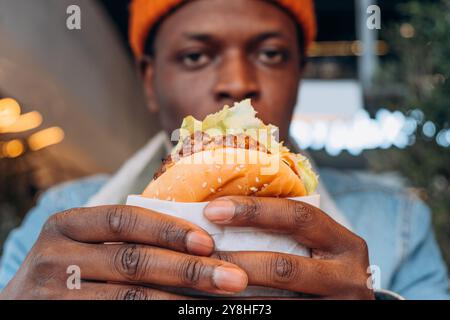 Nahaufnahme eines afroamerikanischen Mannes mit saftigem Burger in der Hand, Fast Food im Restaurant mit köstlichem Hamburger, urbanem Lifestyle. Konzentrieren Sie sich auf das Essen, das Gesicht ist verschwommen Stockfoto