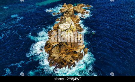 Aus der Vogelperspektive auf eine felsige Insel, umgeben von tiefblauem Meer. Wellen schlagen gegen die Felsen und erzeugen weißen Schaum. Die Insel verfügt über eine zerklüftete Fläche Stockfoto