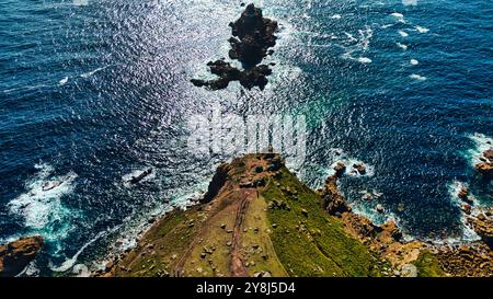 Aus der Vogelperspektive auf eine felsige Küste mit einer kleinen Insel im Meer. Das Wasser schimmert unter Sonnenlicht, was einen schönen Kontrast zum Griee schafft Stockfoto