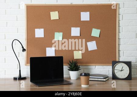 Korkbrett mit leeren Notizen, Laptop, Lampe, Wecker und Notizbüchern auf Holztisch in der Nähe der weißen Ziegelwand Stockfoto