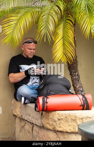 Ein Vielreisender hält an und ruht sich mit seiner Bettrolle und seinem Rucksack auf einer Steinmauer an der State Street in Santa Barbara, Kalifornien aus. Stockfoto
