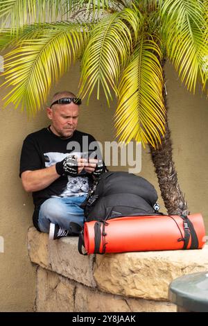 Ein Vielreisender hält an und ruht sich mit seiner Bettrolle und seinem Rucksack auf einer Steinmauer an der State Street in Santa Barbara, Kalifornien aus. Stockfoto